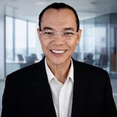 Professional headshot of an Asian man wearing glasses, black blazer, and white shirt, smiling in a modern office setting